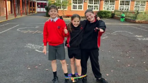 Left to right: Charlie, Evronie and Beau stand with linked arms and the skipping rope around their legs in their playground. They are all smiling. 