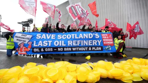 PA Media Unite union members protest - they are holding a banner saying SAVE SCOTLAND'S LAST OIL REFINERY up while waving red flags with slogans on them in the air. A large pile of yellow hard hats is in front of them.