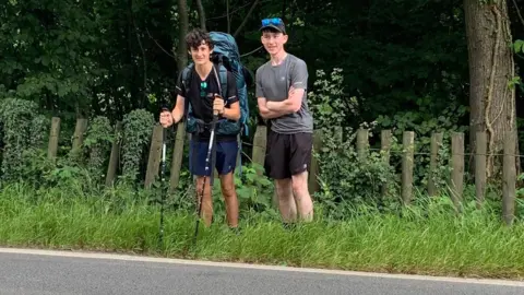 Step2Ukraine James McCarthy-Hill and Adam Mitchell standing on a grassy verge beside the side of a road. They are both wearing shorts and T-shirts. Adam has blue reflective sunglasses resting on his black cap, and James is carrying a large blue backpack and holding two hiking poles. Behind them there is a log and wire fence and thick woodland.