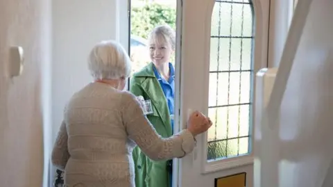 Getty Images An elderly woman with white hair answers the front door to a care worker. The visitor is showing her ID. The walls are white and there is a staircase banister on one side of the hallway.