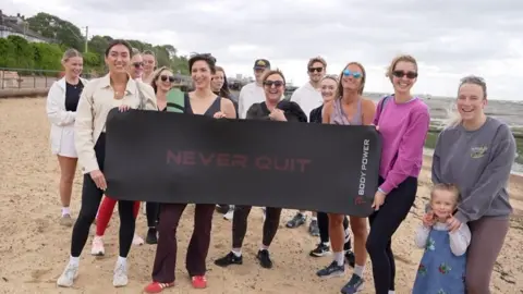 Connor Bennett/BBC About a dozen people on a beach holding a yoga mat that says "don't quit"