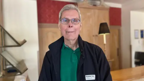 Francis Puddester is an older man with grey hair. He is standing in a hotel reception room with a wooden cabinet and lamp behind him. He is wearing a black jacket and green polo shirt and is smiling into the camera. He has a white paper name badge on.