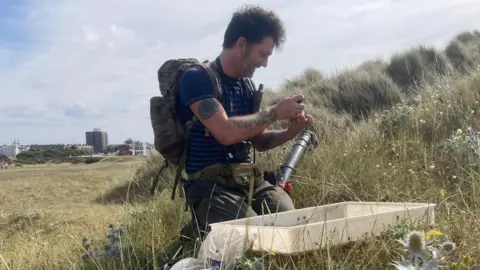 A man in his forties wearing a backpack sits in a field smiling. He has a plastic container and a plastic cylinder next to him and he is examining something in his hand. 