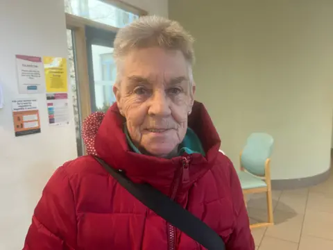 A woman in a red jacket with stands in an NHS building. The floor is tiled and the walls are white and have signs stuck to them. There is a chair in the background. 