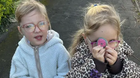 John Devine/BBC Phoebe on the left and Gracie on the right both have long blonde hair and are wearing pink spectacles. Phoebe is wearing a fluffy pale blue coat while Gracie has a leopard print jacket on. Gracie is holding up a pink painted stone to her face.