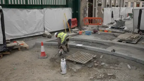 A worker in a hi-vis top with roadworks around them and metal gates with white sheets of plastic.