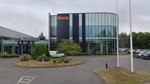 A glass-fronted building behind a mini roundabout with the words, in orange, "Mazak" on the outside. 