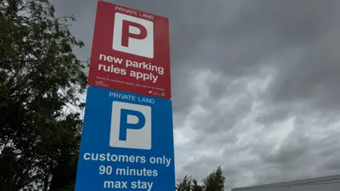 A parking sign outside a Lidl store displays the messages "new parking rules apply" and "customers only, 90 minutes max stay". The sign is pictured against a grey, cloudy sky with part of the supermarket and car park visible below.