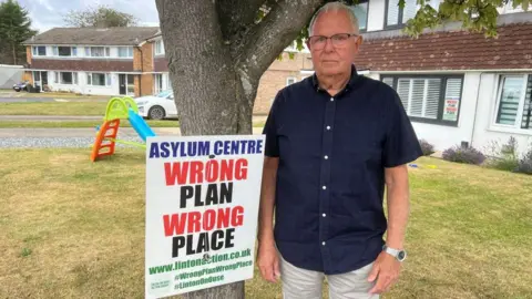 The photograph shows a man wearing a dark short-sleeved shirt, light-coloured shorts and glasses, standing beside a campaign sign attached to a tree on a residential street. Attached to the tree is a printed campaign poster which reads ASYLUM CENTRE WRONG PLAN WRONG PLACE. 