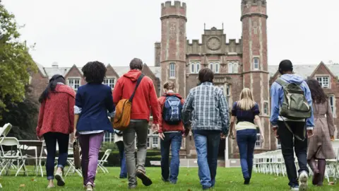 Getty Images A group of eight students are walking towards a university building. They are outside and beside them are plastic chairs 
