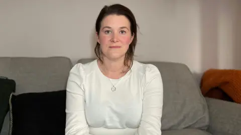 Vicky is wearing a white long-sleeve top and jeans sits on a grey sofa indoors, with cushions and a phone resting beside them.