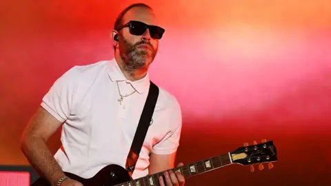 Chase & Status performing on stage at Radio 1's Big Weekend in Luton, with a man on stage playing a black guitar while wearing black sunglasses and a white tshirt. The background is lit up in orange and pink.