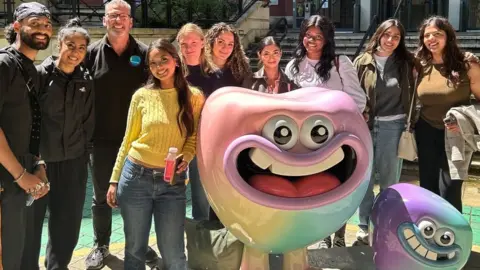 Phil Loughnane Phil and about nine students are standing outside the dental school posed around two sculptures of teeth with faces showing large cartoon-style eyes and wide-open mouths. They are all smiling to camera in sunshine 