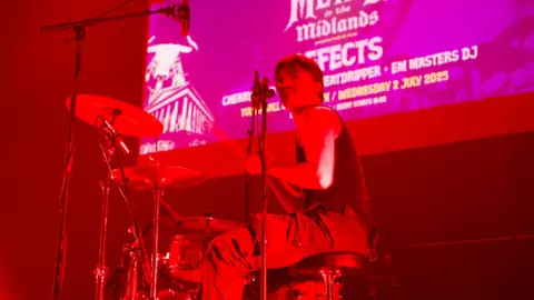 A man in a black vest playing drums on stage