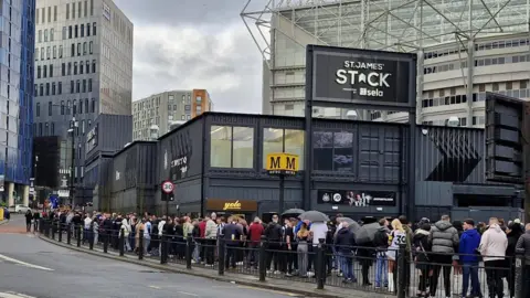 Una fila de personas paradas fuera de la pila de St. James está compuesta por contenedores de envío negro.