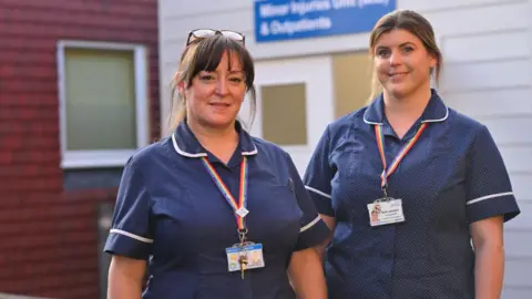 Two female nurses stand smiling outside a hospital building. 