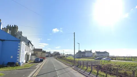 Google A screenshot from Google Streetview showing the main road through Allonby. There are terraced houses on the left, with a large open space on the right, where some cars are parked and there are some houses and a play park in the distance. 