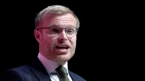Getty Images Michael Shanks has fair hair and beard, glasses and a dark blue suit over a white shirt and a dark green tie. He speaking is in an indoor space.