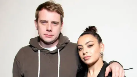 Getty Images A man with light brown wearing a brown hoodie with white drawstrings and a white T-shirt underneath, looks into the camera. He has his left arm around a woman wearing her dark hair in a tight bun. She is wearing a dark top and large earrings.