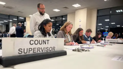 Emma Baugh/ BBC People sit in a row at a table counting ballot papers. A man stands behind them overseeing their work. The words 'Count Supervisor' is on a display to the left.