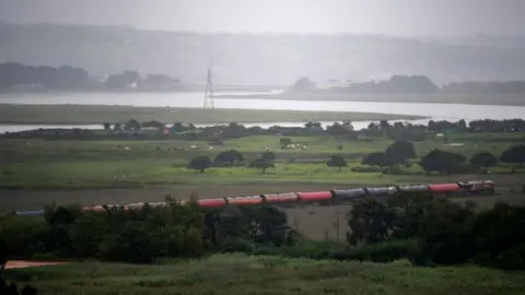 Getty Images A train travelling through a rural area with a river and marshes seen in the background.