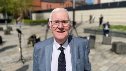BBC Eugene Reavey has short grey hair and wire framed glasses and is wearing a checked blue/grey suit, white shirt and dark spotted tie