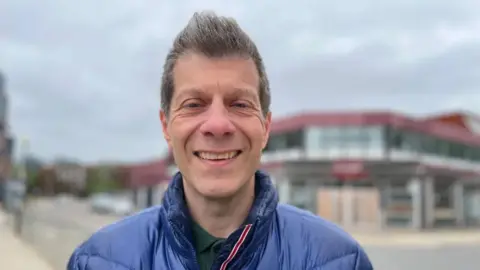 Emma Baugh/BBC Pep, a man, wearing a blue jacket, smiling for the camera. He is outside and the background is out of focus. It is cloudy above.