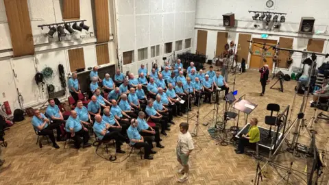 A full choir, pictured from above as they record their voices. Each is wearing a light blue polo shirt and dark trousers. Microphones and recording equipment can be seen in front of them.