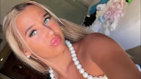 A teenage girl with blonde hair and a white top looks at the camera