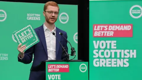 Ross Greer, who has short ginger hair, holds a green manifesto in his right hand while speaking into a podium. He is wearing a blue suit and white shirt, with green branding behind him 
