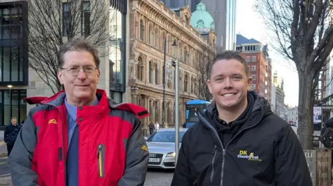 Two men smiling at the camera. They are on a busy city centre street (Bedford Street in Belfast) with cars, buses and pedestrians visible in the background. Jonathan is wearing a red coat with grey sleeves over a blue jumper, he has brown hair and round glasses. Dale is younger and has a black jacket on over a black hoody. He has short brown spiky hair.