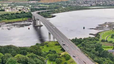 An aerial view of Foyle Bridge. A large grey bridge between two grassy land masses. A number of cars are visible crossing the bridge over the River Foyle. 