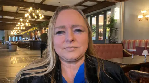 BBC Shows a woman in blond hair with a blue blouse and black jacket standing in a hotel lobby