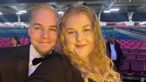 PA Media Ellis Pears wearing a tuxedo, with a white shirt and black bow tie. He is stood next to a woman with long wavy blonde hair. There are rows of seats behind them in a hall.