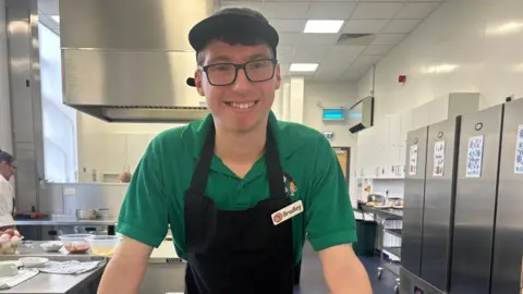 BBC Bradley stands in the kitchen and wears a green T-shirt, black apron with his name badge on it and a cap that covers his short dark hair. He is wearing glasses and is smiling.