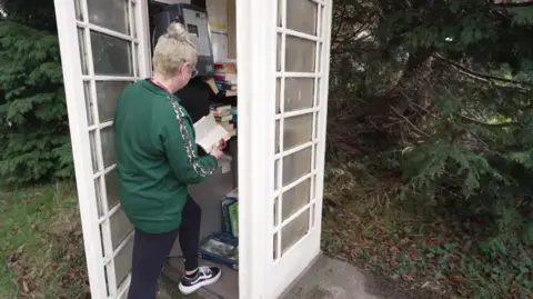 BBC A woman reads a book as she stands in the doorway of a mid-20th century cream-coloured telephone box. More books can be seen stacked inside, alongside a pay phone. The woman has blond hair, tied in a bun, and wears a green top and black leggings.