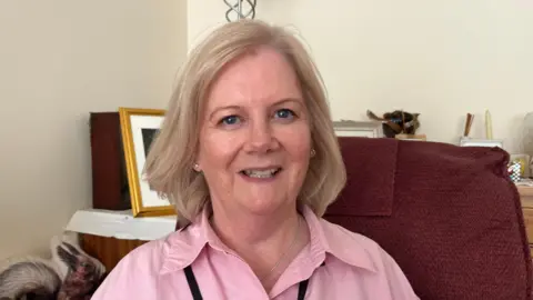 A woman looking into the camera smiling. She has blonde hair in a bob and is wearing a pink shirt with the top button open. She is sitting on a red armchair in a loving room - there is a table with a gold picture frame behind her. We cannot see the photo inside.