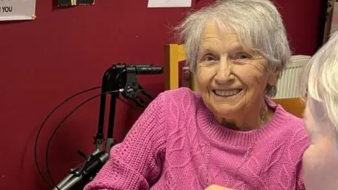 Pensioner Mary has grey hair and is wearing a pink jumper. She is smiling at the camera