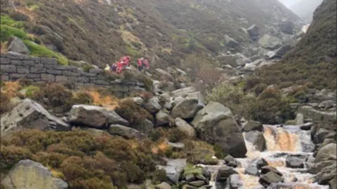 In the distance mountain rescuers, all dressed in red, help a family across a fast flowing river as water cascades down rocks