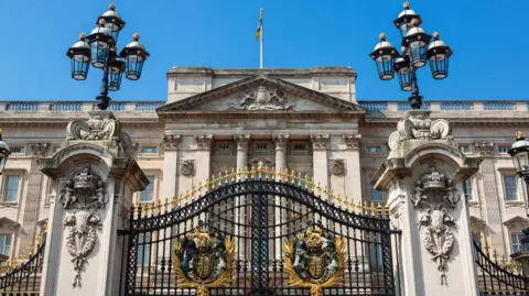 Getty Images Exterior of Buckingham Palace. It is simply a ample  stone-coloured gathering  with columns and a balustrade astatine  the top. In the foreground, determination   are 2  ample  pillars with lanterns connected  apical  of them and ornate achromatic  and golden  gates successful  the middle. 