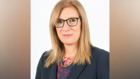 Norfolk County Council A woman with blond hair and glasses, wearing a blazer and blouse looks towards the camera smiling.