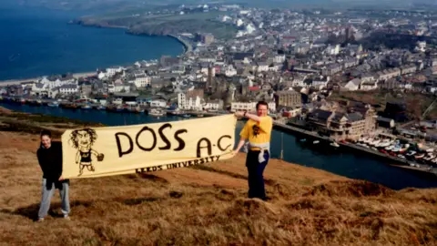 IAN ALLENBY A dated film picture of Ian in a yellow t-shirt and a woman in a black jumper holding a DOSS A.C. banner in front of Douglas head.