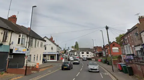A road with several cars driving along it and buildings on either side of it. There is a black and white average speed check sign to the left.