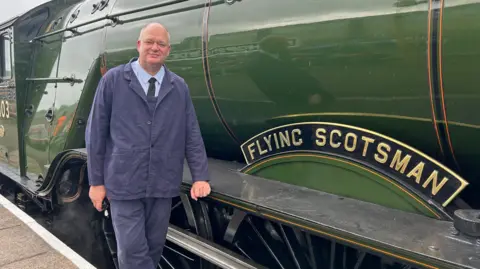 A man in a blue suit is standing next to a locomotive which says Flying Scotsman on it. It is dark green.