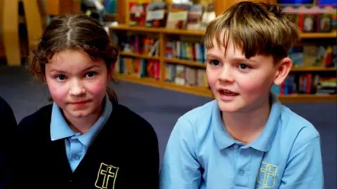 Two school pupils, a boy and a girl sit in the school library in their light blue uniforms.