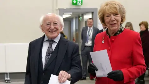 PA Michael D Higgins in black suit and white shirt, wife Sabina in red coat and black gloves, holding ballot papers and voting