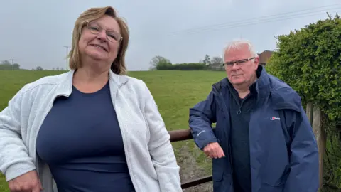 Patrina Needham, with shoulder-length brown hair and classes, leans on a metal gate with a field behind it, next to Mark Benzing who has short white hair, glasses and is wearing a large dark blue waterproof jacket. Both pose for a photo - Patrina is smiling and Mark looks more serious. 