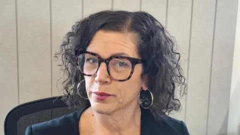 Leicestershire Police Nicola Potts headshot, showing a woman with mid length wavy black hair, hoop earrings and spectacles sat on an office chair in front of a window