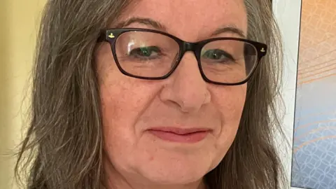 A close-up photo of a long haired woman with black glasses smiling at the camera.