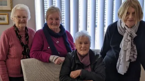 Wincanton Cares Four women are smiling at the camera. Three are standing and one lady is sitting in a chair. There are pictures on the wall behind them along with a window with blinds.
